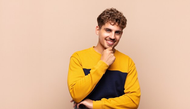 Joven sonriendo, disfrutando de la vida, sintiéndose feliz, amigable, satisfecho y despreocupado con la mano en la barbilla