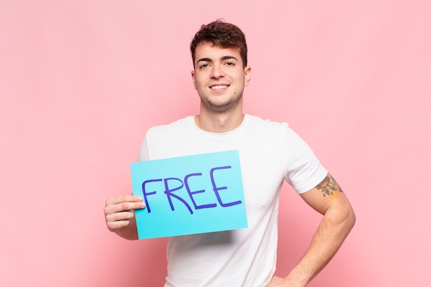 Joven sonriendo felizmente con una mano en la cadera y actitud confiada, positiva, orgullosa y amistosa