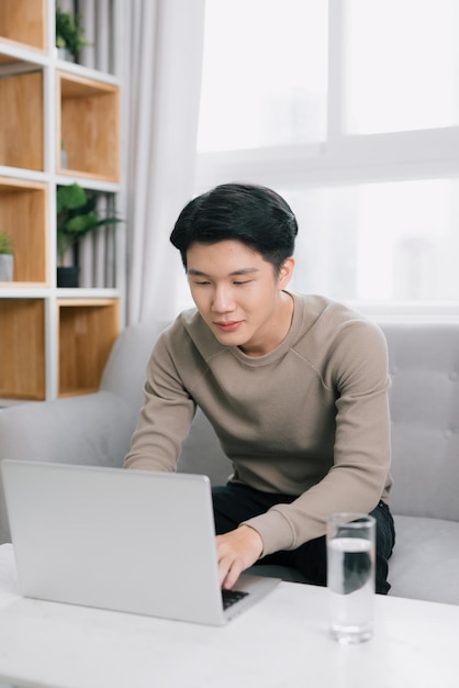 Joven sonriendo mientras lee la pantalla de una computadora portátil mientras se relaja en un cómodo sofá en casa