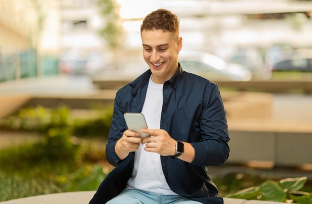 Un joven sonriendo mientras mira su teléfono inteligente sentado al aire libre en un banco