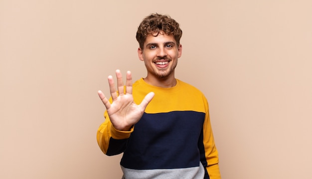 Joven sonriendo y mirando amigable, mostrando el número cinco o quinto con la mano hacia adelante, contando hacia atrás