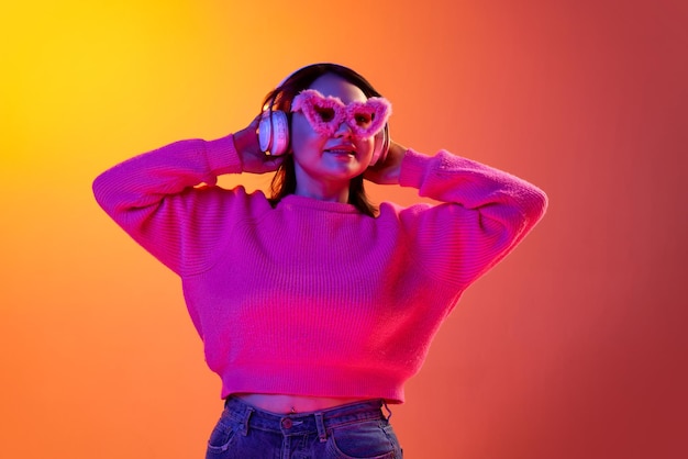 Joven sonriente con acogedoras gafas de piel de suéter escuchando música en auriculares sobre fondo naranja degradado en luz de neón Concepto de emociones estilo de vida juvenil