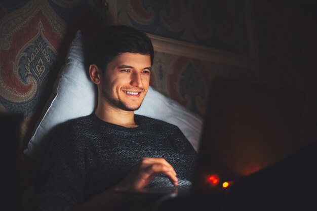 joven sonriente acostado en la cama con el portátil en una habitación oscura en casa.