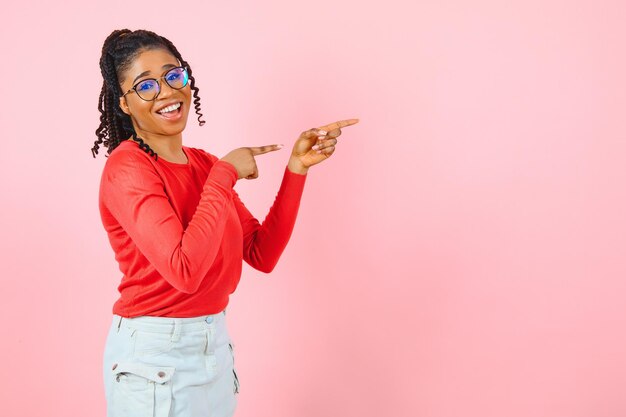 Joven sonriente alegre mujer afroamericana cabello rizado en suéter de punto casual apuntar los dedos índices a un lado en el espacio de trabajo del espacio de copia simulacro aislado en un retrato de estudio de fondo rosa pastel.