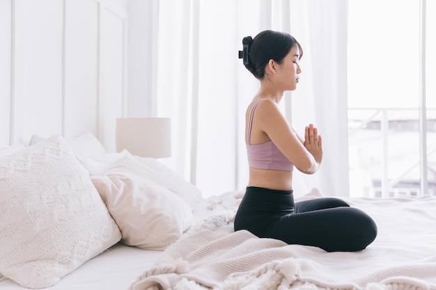 Joven sonriente y atractiva mujer asiática deportiva practicando yoga en la cama haciendo ejercicio Ardha Padmasana meditando en la pose de Half Lotus con namaste haciendo ejercicio en casa usando ropa deportiva
