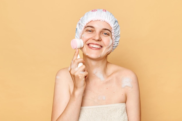 Joven sonriente y atractiva mujer limpiando la cara con un cepillo de pie usando gorro de baño expresando felicidad disfrutando de un tratamiento de belleza de pie aislado sobre un fondo beige