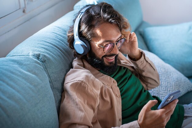 Un joven sonriente con auriculares escucha música en su teléfono inteligente sentado en el sofá de su casa