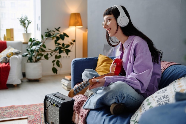 Joven sonriente con auriculares sentada en el sofá y aprendiendo a tocar la guitarra