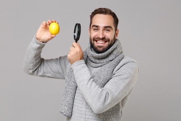 Joven sonriente con bufanda de suéter gris aislado en un retrato de estudio de fondo gris. Estilo de vida saludable tratamiento de enfermedades enfermas, concepto de temporada fría. Simulacros de espacio de copia. Sostenga el limón de la lupa.