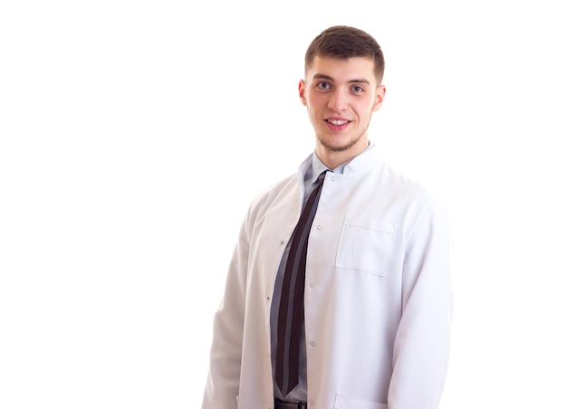 Joven sonriente con cabello castaño en camisa azul, corbata y bata de médico sobre fondo blanco en el estudio