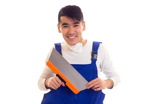 Joven sonriente con cabello oscuro en camisa blanca y azul en general con respirador blanco sosteniendo una espátula sobre fondo blanco en el estudio