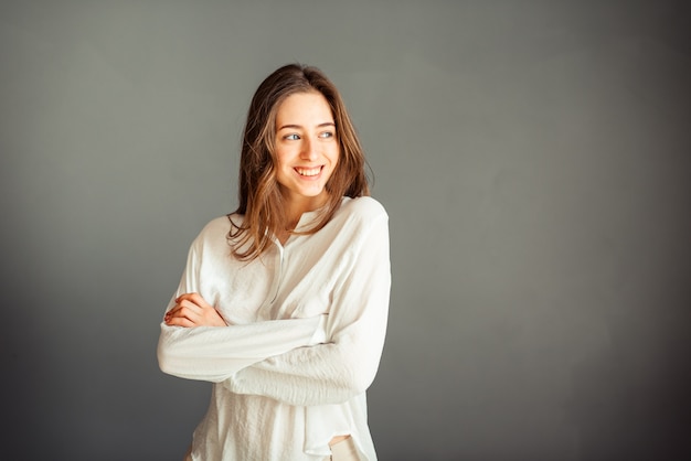 Joven sonriente con una camisa blanca, la cabeza inclinada hacia un lado, la mano cerca de la mejilla, la segunda en el bolsillo, el aislante en una pared gris. Sin retoques. Sin maquillaje.