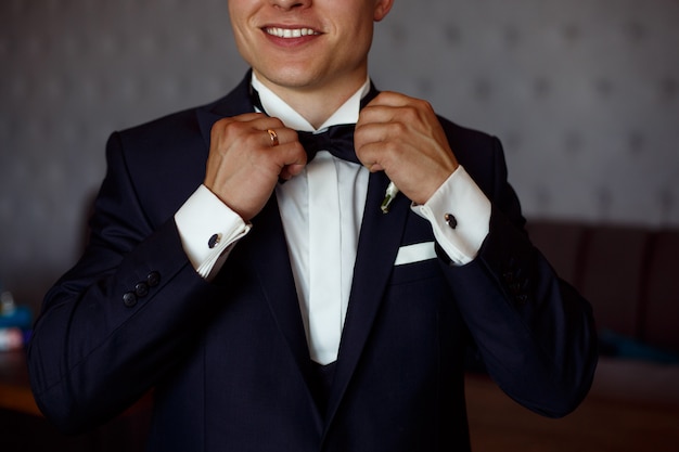 Foto joven sonriente en camisa blanca y traje negro ajusta la mariposa de cerca. chico elegante pone un traje de negocios. novios mañana cerca