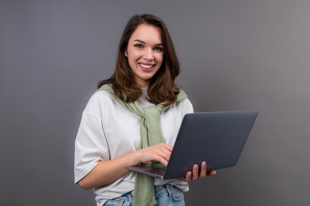 Una joven sonriente con una camiseta blanca y un suéter se para con una computadora portátil en un fondo gris y mira a la cámara Retrato de un profesional independiente