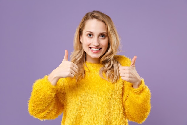 Una joven sonriente y divertida con suéter de piel amarilla posando aislada en un retrato de estudio de fondo de pared violeta pastel. Gente emociones sinceras concepto de estilo de vida. Simulacros de espacio de copia. Mostrando los pulgares hacia arriba.
