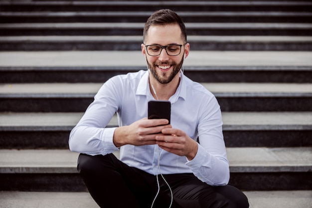 Joven sonriente elegantemente vestido sentado en las escaleras con auriculares en los oídos y usando teléfonos inteligentes para revisar mensajes en las redes sociales.