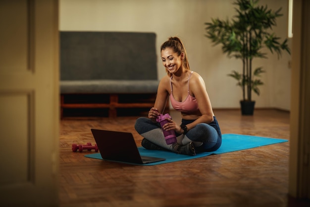 Una joven sonriente está viendo tutoriales en Internet y entrenando deportes en la sala de estar en la alfombra del piso frente a la computadora portátil.