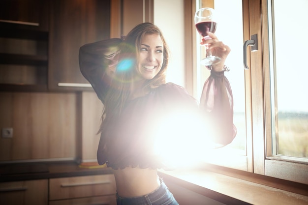 Foto una joven sonriente feliz está bebiendo vino tinto y brindando por alguien, mientras está de pie junto a la ventana.