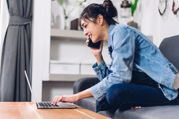 Joven sonriente feliz hermosa mujer asiática relajante usando la computadora portátil trabajando y reunión de video conferencia en casa. Joven charla creativa con el teléfono inteligente. Trabajar desde el concepto de hogar