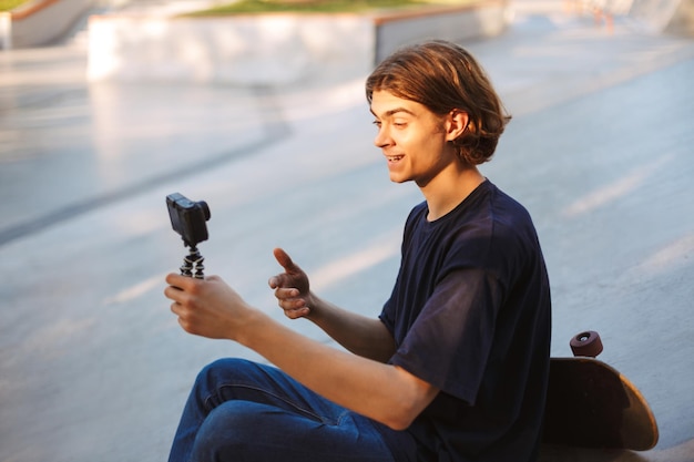 Joven sonriente grabando felizmente un nuevo video para vlog con patineta cerca del skatepark moderno