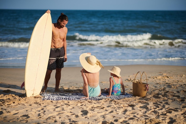 Joven sonriente hablando con su esposa y su pequeña hija después de surfear en el mar