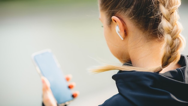 Joven sonriente haciendo deporte y corriendo en el parque usando su teléfono para escuchar música con auriculares inalámbricos al atardecer en la ciudad mirando la pantalla