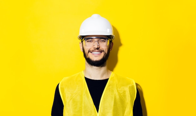 joven sonriente, ingeniero constructor, con casco de seguridad de construcción blanco, gafas y chaqueta amarilla aislada en la pared amarilla.