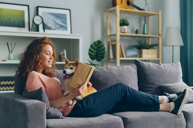 Una joven sonriente leyendo un libro en casa en el sofá y abrazando a un lindo perro mascota