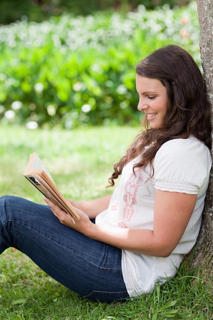 Joven sonriente, leyendo un libro mientras se inclina contra un árbol