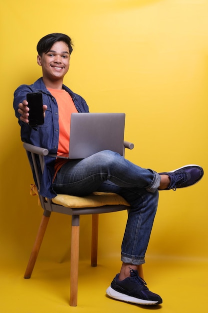 Un joven sonriente mostrando un teléfono móvil con pantalla en blanco a la cámara para simular mientras usa una computadora portátil y ove
