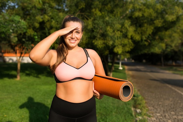 Foto joven y sonriente mujer de talla grande con top deportivo rosa sosteniendo una alfombra de yoga naranja en la mano mientras mira alegremente a la cámara pasando tiempo en el parque de la ciudad