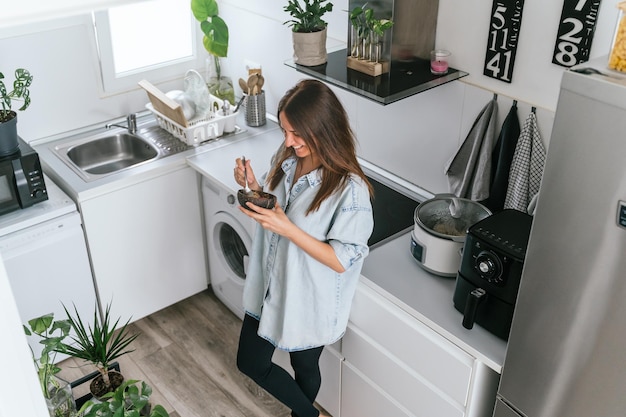 Una joven sonriente parada en la cocina de casa tomando un refrigerio o desayunando