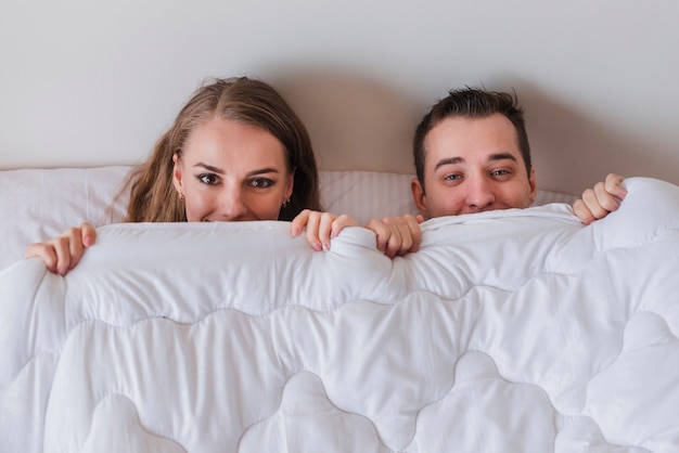 Joven sonriente pareja acostada en la cama y mirando fuera de edredón