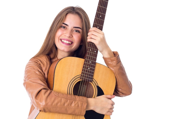 Una joven sonriente con el pelo largo y una chaqueta marrón abrazando una guitarra con fondo blanco en el estudio