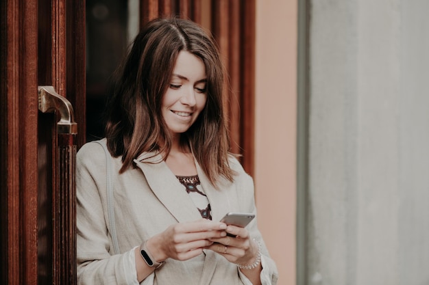 Una joven sonriente con el pelo oscuro vestido con una elegante chaqueta blanca sostiene modernos puestos de teléfonos inteligentes cerca de las puertas de los mensajes del edificio de oficinas con amigos en las redes sociales Concepto de tecnología