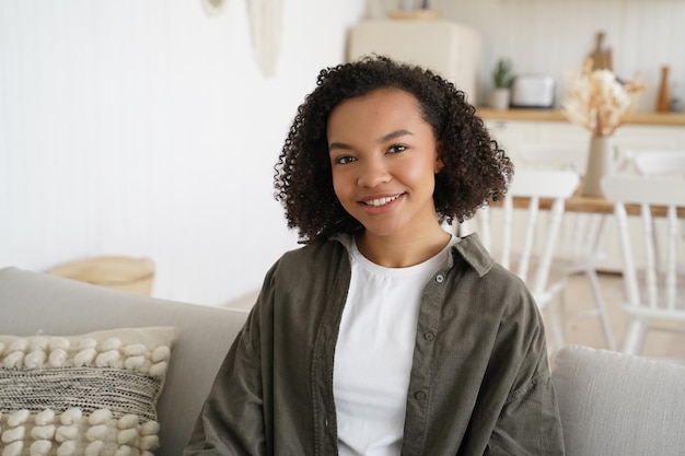 Una joven sonriente de raza mixta con peinado afro se sienta en un sofá en una acogedora sala de estar en casa