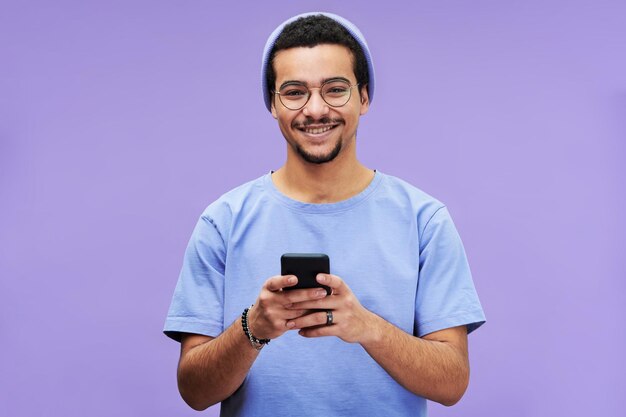 Joven sonriente con ropa casual azul mirando a la cámara mientras usa el teléfono inteligente