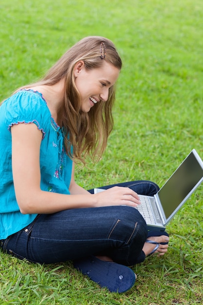 Joven sonriente, sentado con las piernas cruzadas en el campo con su computadora portátil