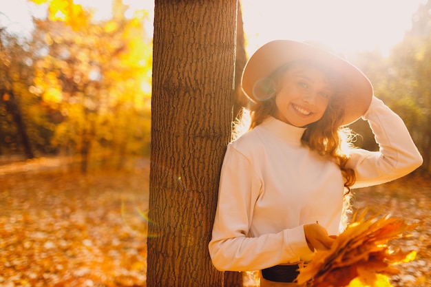 Una joven sonriente con sombrero disfruta del clima otoñal en el bosque con las hojas amarillas al atardecer