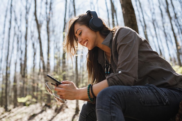 Una joven sonriente usando su teléfono inteligente y escuchando música mientras disfruta en el bosque.