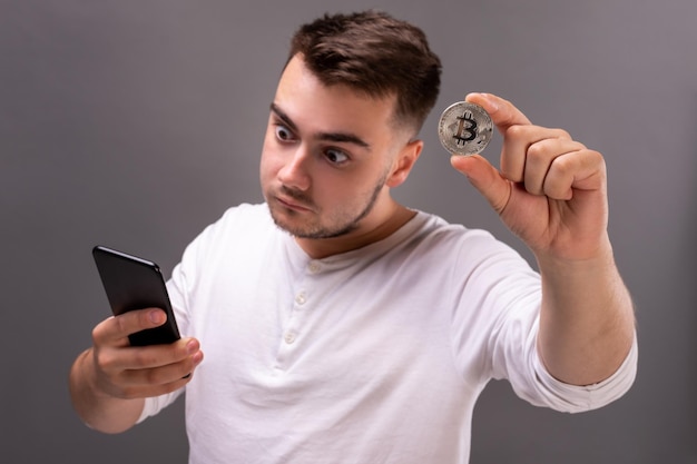 El joven se sorprende al ver el mercado de criptomonedas en su teléfono. Sosteniendo una moneda de bitcoin en su mano