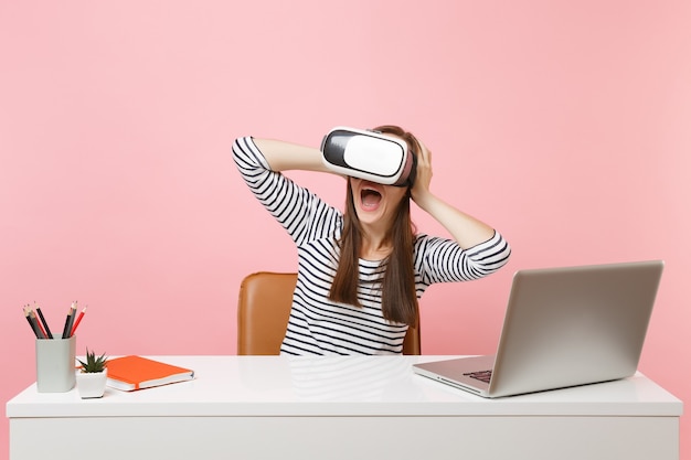 Joven sorprendida en auriculares de realidad virtual aferrándose a la cabeza gritando sentarse y trabajar en un escritorio blanco con un portátil aislado sobre fondo rosa pastel. Concepto de carrera empresarial de logro. Copie el espacio.