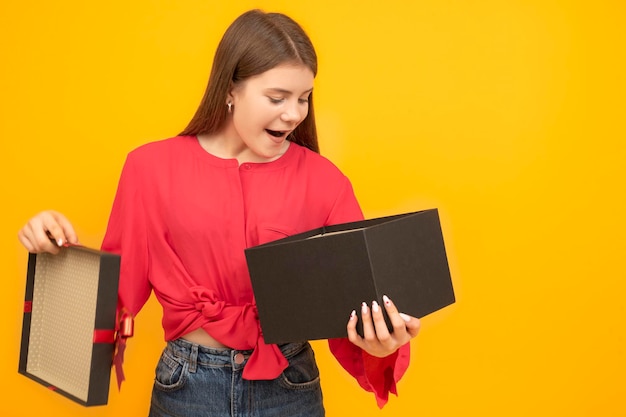 Una joven sorprendida y entusiasta sostiene un regalo abierto Mujer con una gran caja negra sobre fondo amarillo brillante