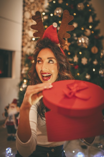 Una joven sorprendida está sentada en el suelo junto al árbol de Navidad y sostiene el regalo mágico en la caja roja que acaba de recibir.