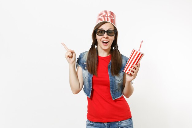 Joven sorprendida en gafas 3d con cubo para palomitas de maíz en la cabeza viendo una película sosteniendo una taza de refresco o cola apuntando con el dedo índice en el espacio de la copia aislado sobre fondo blanco. Emociones en el cine