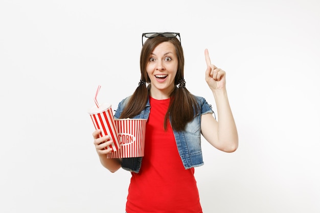 Joven sorprendida en gafas 3d viendo una película, sosteniendo un cubo de palomitas de maíz, un vaso de plástico de refresco o cola apuntando con el dedo índice hacia arriba en el espacio de la copia aislado sobre fondo blanco. Emociones en el cine.