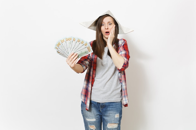 Joven sorprendida hermosa mujer con sombrero de periódico sosteniendo un paquete de dólares, dinero en efectivo y aferrándose a la cara aislada sobre fondo blanco. Instrumentos para la renovación de la habitación del apartamento. Reparar el concepto de hogar.