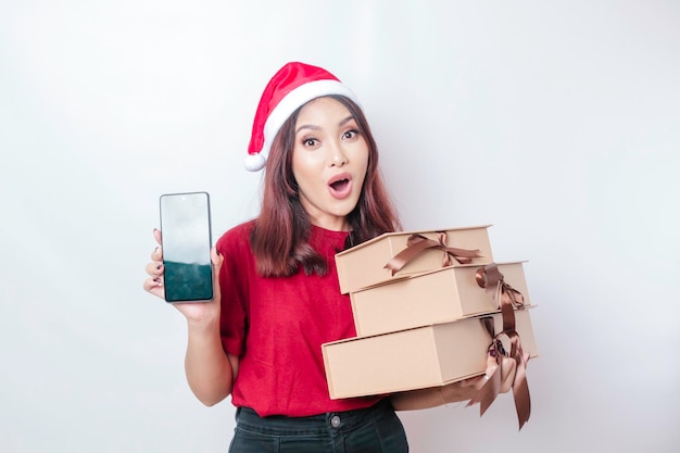 Una joven sorprendida lleva un sombrero de Papá Noel mientras sostiene regalos de Navidad y su teléfono inteligente