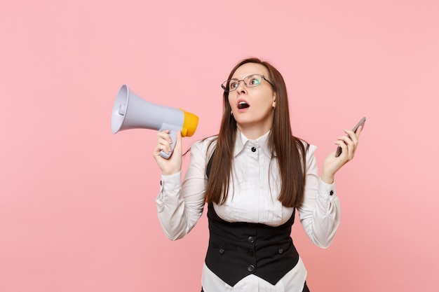 Joven sorprendida mujer de negocios agotada en traje, gafas mirando hacia arriba sosteniendo megáfono y teléfono móvil aislado sobre fondo rosa. Jefa. Riqueza de carrera de logro. Copie el espacio para publicidad.