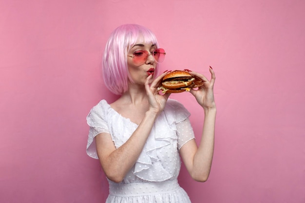 Joven sorprendida con el pelo corto rosa sostiene una hamburguesa y mira una hamburguesa